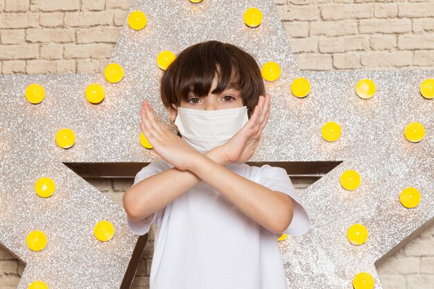 A front view cute little kid in white t-shirt dark jeans white sterile mask on the star designed yellow stand and light background