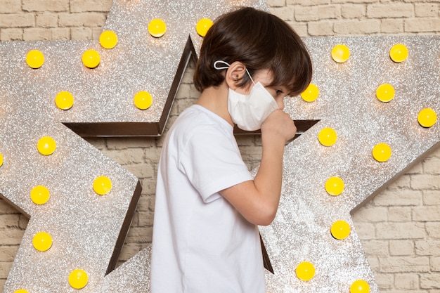 A front view cute little kid in white t-shirt dark jeans white sterile mask on the star designed yellow stand and light background