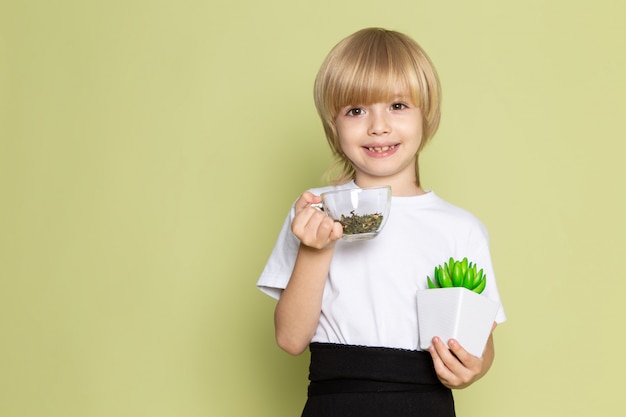 Free photo a front view cute smiling kid in white t-shirt holding species and green little plant on the stone colored desk