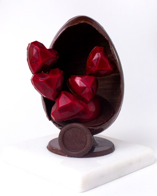 Front view decorated chocolate egg with chocolate red candies in the shape of a heart inside on stand