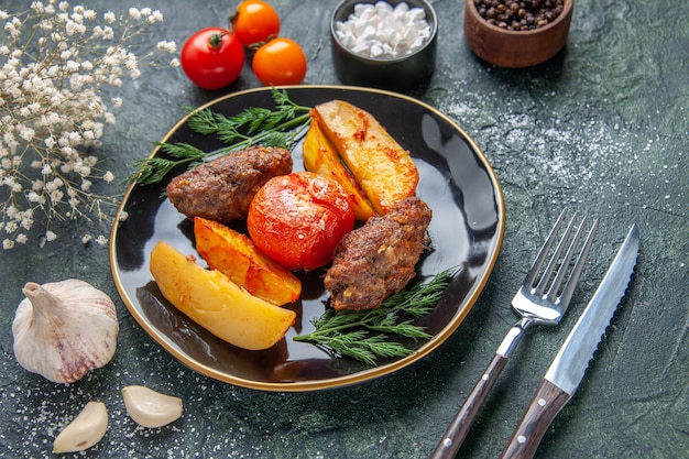 Free Photo front view of delicious meat cutlets baked with potatoes and tomatoes on a black plate cutlery set white flowers spices garlics on green black mixed colors background