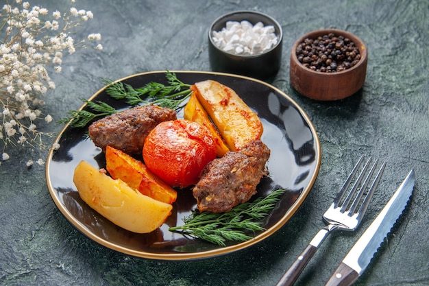 Free photo front view of delicious meat cutlets baked with potatoes and tomatoes on a black plate cutlery set white flowers spices on green black mixed colors background