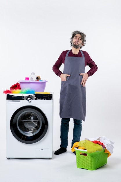 Front view determined housekeeper man putting hand in pocket standing near white washing machine on white background