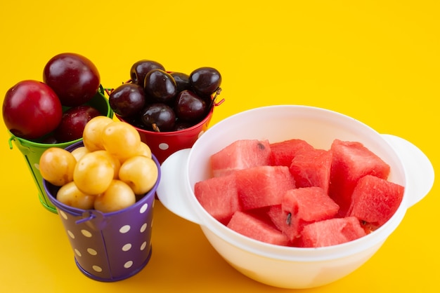 A front view different fruits inside baskets with sliced watermelon on yellow, color fruit composition