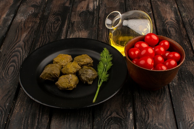 front view dolma inside black plate along with fresh red cherry tomatoes and olive oil on the brown wooden rustic floor