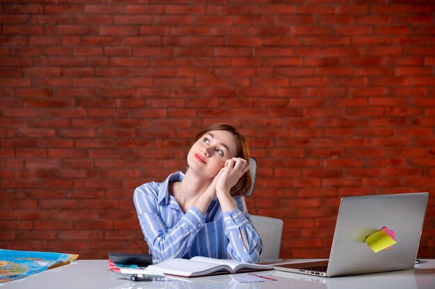 Front view dreaming travel agent sitting behind her working place with laptop agency service assistant ticket manager map operator global world