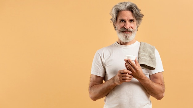 Front view of elder bearded man holding moisturizer with copy spadce