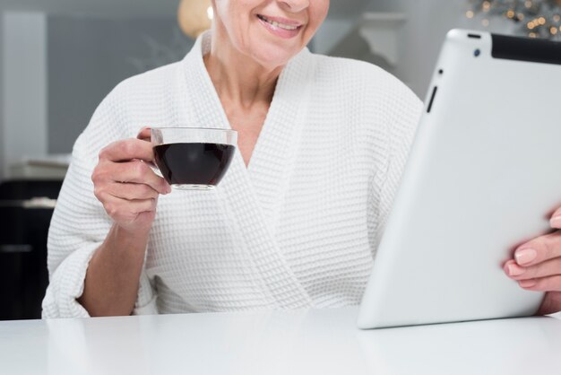 Front view of elder woman in bathrobe holding tablet and coffee cup