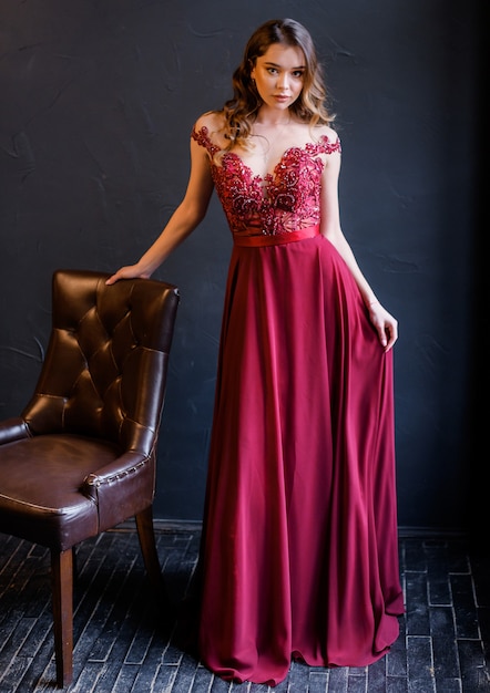 Free photo front view of elegant girl in a red dress leans on a chair and looks at the camera