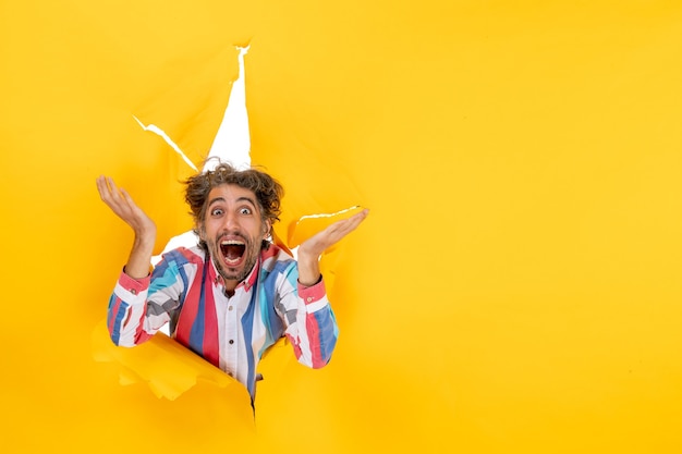 Front view of an emotional and crazy young guy smiling through a torn hole in yellow paper