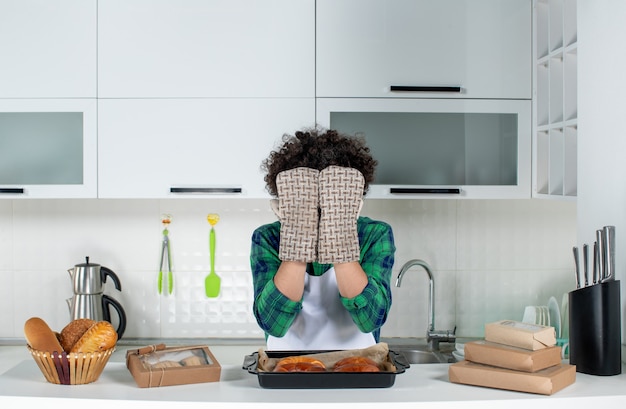 Free Photo front view of emotional guy wearing holder covering his face standing behind table with freshly-baked pastry on it in the white kitchen