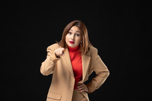 Front view of emotional young lady in a light brown suit pointing someone on dark