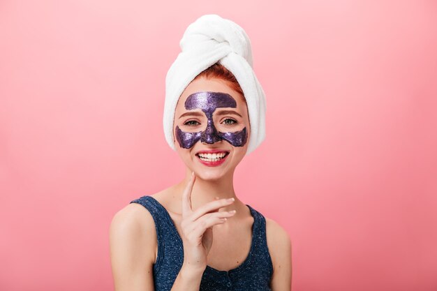 Front view of enchanting woman with face mask laughing on pink background. Studio shot of blissful girl with towel on head doing spa treatment.