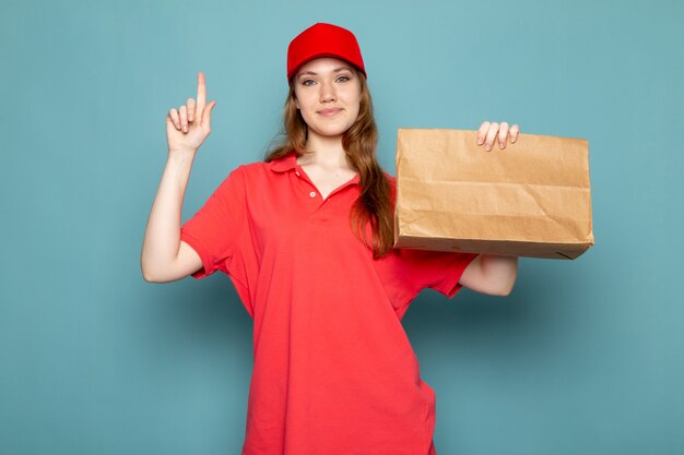 A front view female attractive courier in red polo shirt red cap holding brown package smiling on the blue background food service job