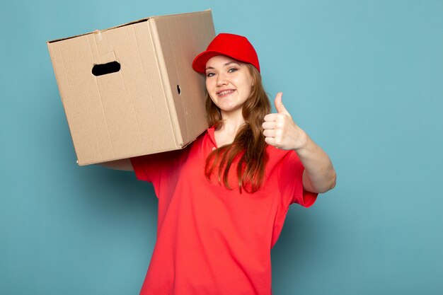 A front view female attractive courier in red polo shirt red cap and jeans holding box posing smiling on the blue background food service job