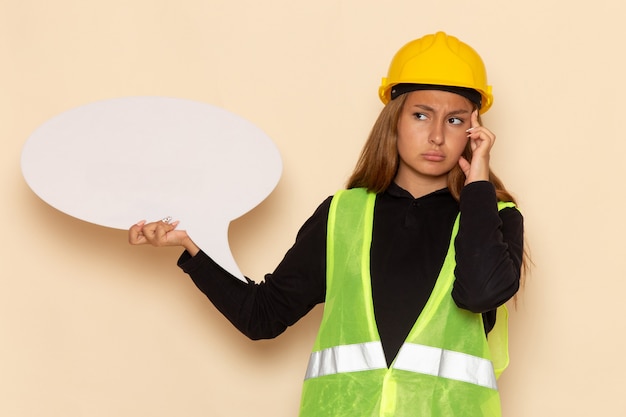 Front view female builder in yellow helmet holding a big white sign and thinking on white wall female  architect