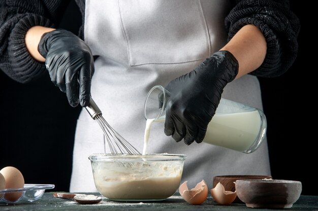 Front view female cook pouring milk into eggs and sugar for dough on dark hotcake pastry cake pie cuisine job worker