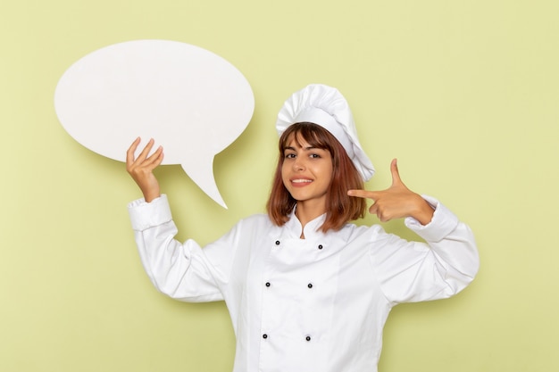 Front view female cook in white cook suit holding a big white sign on a green surface