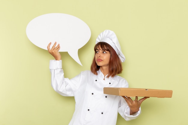 Free photo front view female cook in white cook suit holding food box and white sign on a green desk