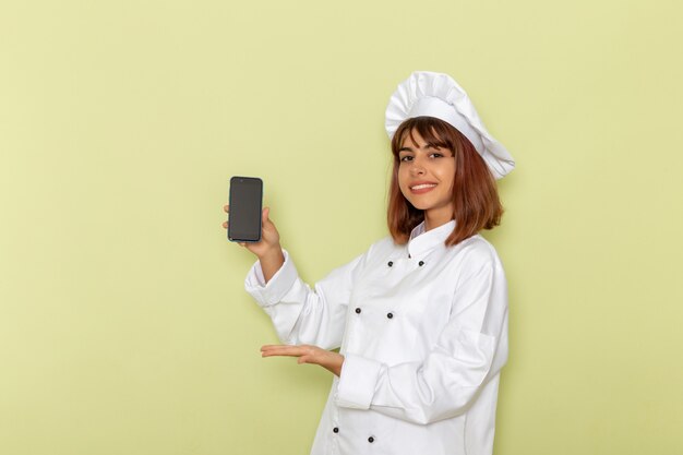 Front view female cook in white cook suit holding her smartphone on a green surface