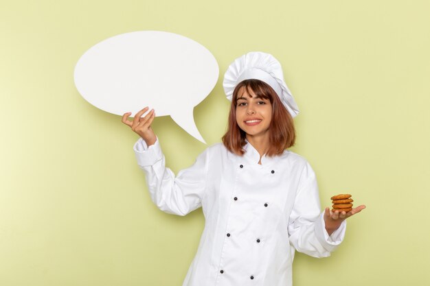 Front view female cook in white cook suit holding little cookies on green surface