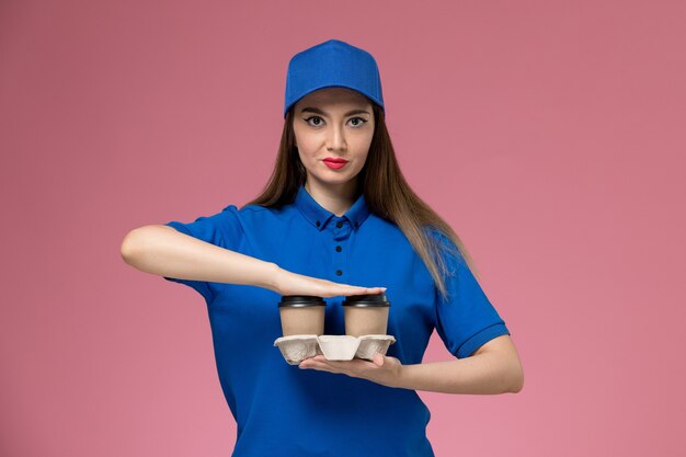 Front view female courier in blue uniform and cape holding brown delivery coffee cups posing on pink wall 