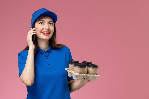 Front view female courier in blue uniform and cape holding coffee cups and talking on the phone on the pink wall 