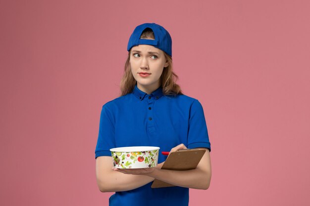 Free Photo front view female courier in blue uniform cape holding delivery bowl and notepad on light pink wall, service delivery job work employee