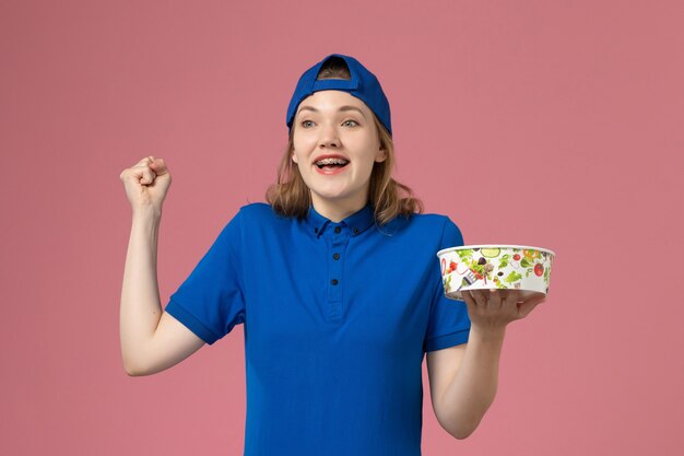 Front view female courier in blue uniform cape holding delivery bowl on pink wall, worker service delivery employee worker job