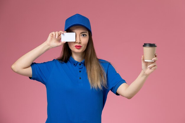 Front view female courier in blue uniform and cape holding delivery coffee cup and white card on pink desk job