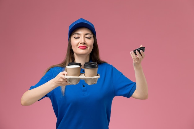 Front view female courier in blue uniform and cape holding delivery coffee cups smelling scent on pink wall 