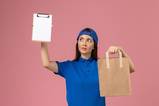 Front view female courier in blue uniform cape holding delivery paper package and notepad on the pink wall, job service employee delivering