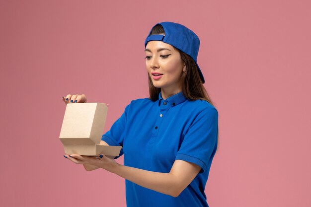 Front view female courier in blue uniform cape holding empty little delivery package on pink wall, worker employee service company delivery