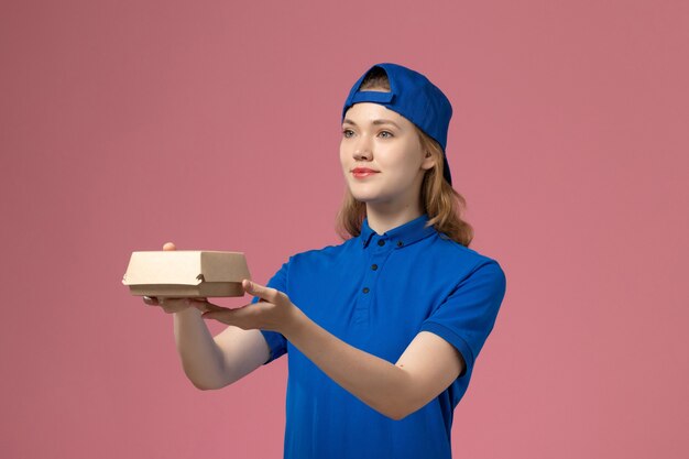 Front view female courier in blue uniform and cape holding little delivery food package on the pink background delivery uniform service company work worker girl