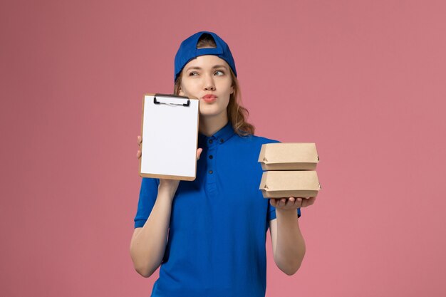 Front view female courier in blue uniform cape holding little delivery food packages and notepad thinking on pink background delivery service employee