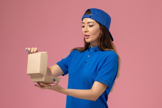 Front view female courier in blue uniform cape holding little delivery package opening it on pink wall, employee service company delivery