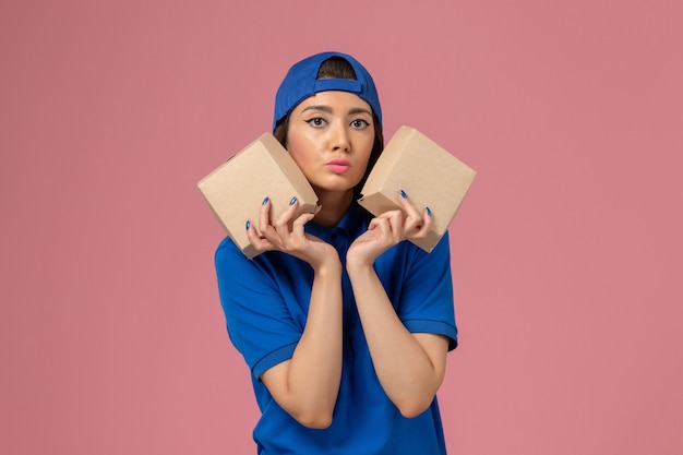 Front view female courier in blue uniform cape holding little delivery packages on light pink wall, service employee delivery