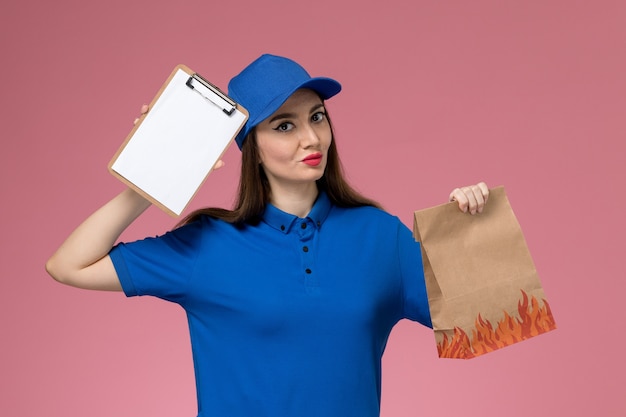 Front view female courier in blue uniform and cape holding notepad and paper food package thinking on pink wall 