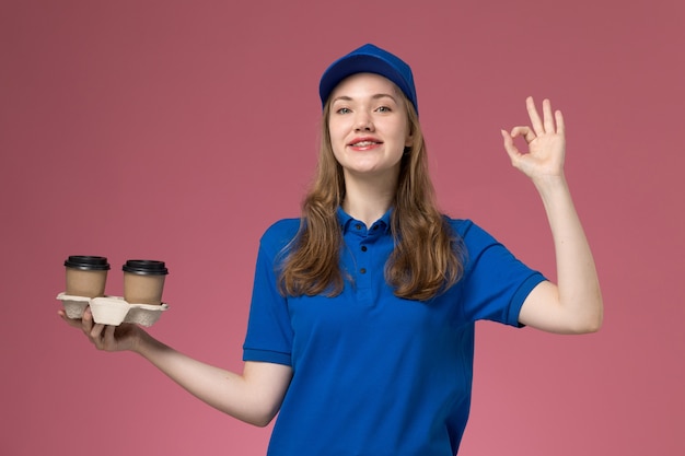 Front view female courier in blue uniform holding brown delivery coffee cups smiling on the pink background service uniform delivering company job