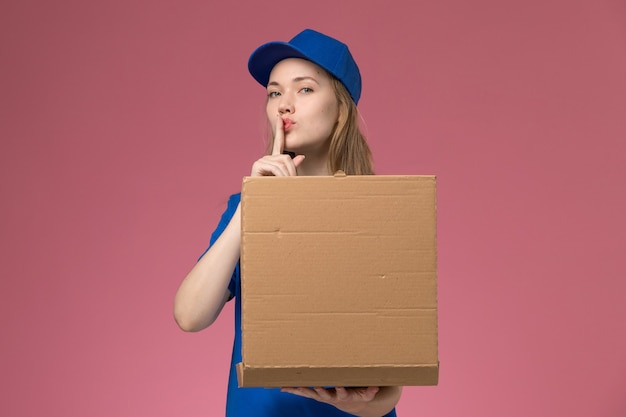 Front view female courier in blue uniform holding food delivery box showing silence sign on pink desk job service uniform company