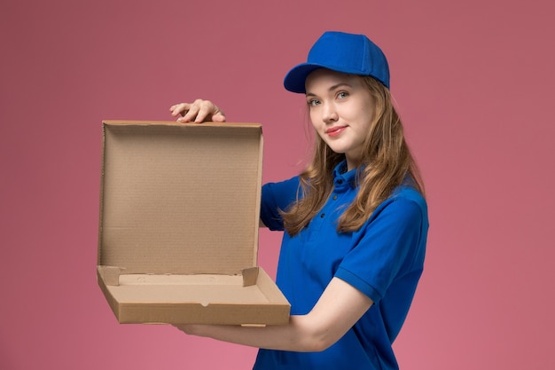 Front view female courier in blue uniform holding open and empty food box on the pink background job worker service uniform company