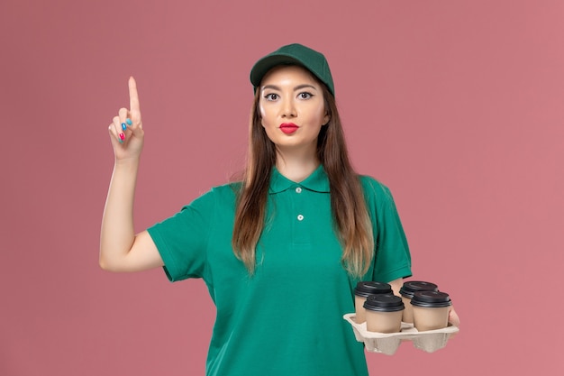 Front view female courier in green uniform and cape holding brown delivery coffee cups on the pink wall service uniform delivery job