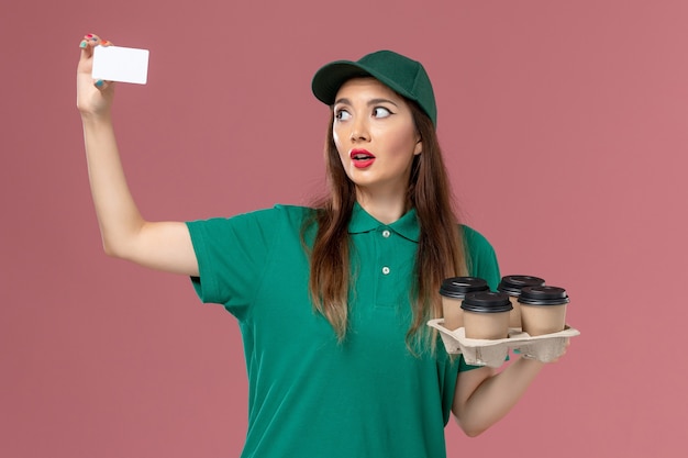 Free photo front view female courier in green uniform and cape holding card and delivery coffee cups on pink wall service uniform delivery job work worker