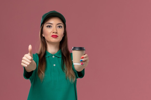 Front view female courier in green uniform and cape holding delivery coffee cup on light-pink wall service job uniform delivery