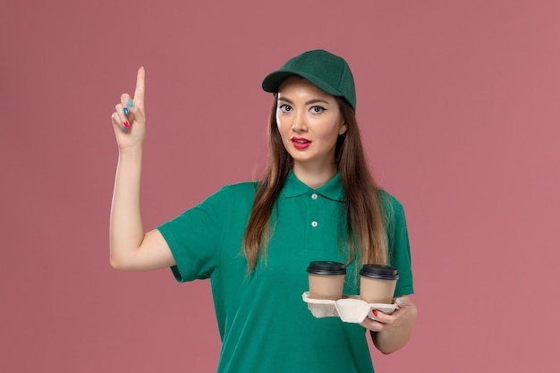 Front view female courier in green uniform and cape holding delivery coffee cups on pink wall company service job uniform delivery worker female work girl