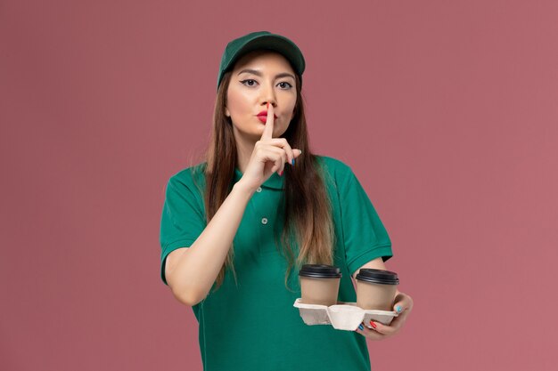 Front view female courier in green uniform and cape holding delivery coffee cups on pink wall company service uniform delivery female work girl