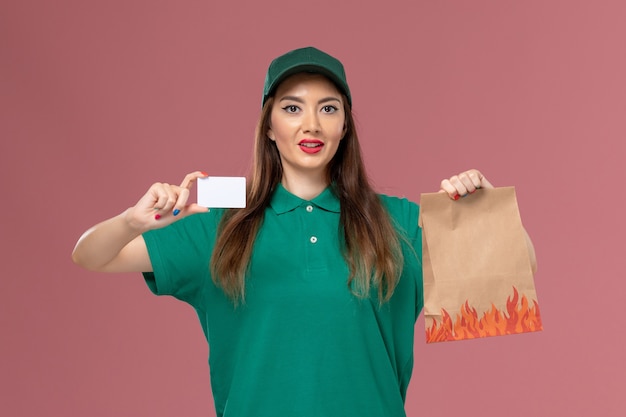 Front view female courier in green uniform holding white card and food package on pink wall service uniform delivery job