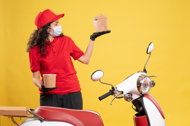 Front view female courier in mask holding delivery dessert and food on yellow background covid- job uniform worker service delivery