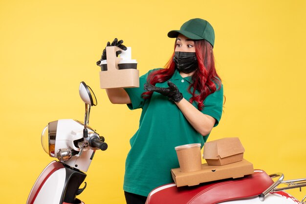 Front view of female courier in mask with delivery coffee on yellow wall