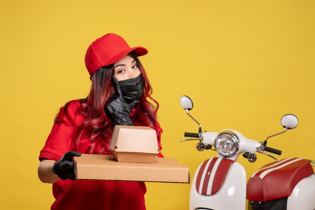 Front view of female courier in mask with delivery food on yellow wall